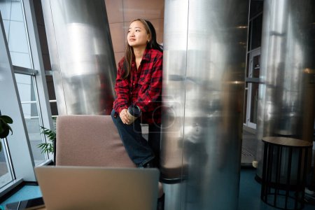 Téléchargez les photos : Jeune asiatique concentrée employée de l'informatique avec tasse de café assis sur la chaise et regardant loin dans le bureau de coworking. Concept de travail indépendant ou à distance moderne - en image libre de droit