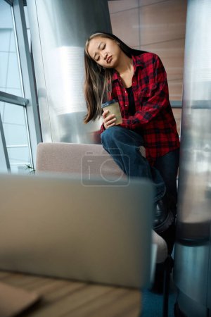 Foto de Joven asiática trabajadora de TI con taza de café sentada en la silla y pensando en algo en la oficina de coworking. Concepto de trabajo independiente o remoto moderno - Imagen libre de derechos