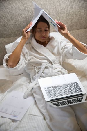 Photo for Top view of young exhausted beautiful caucasian businesswoman wearing bathrobe with laptop covering her head with papers on bed in hotel room at morning time. Business trip, vacation and travelling - Royalty Free Image