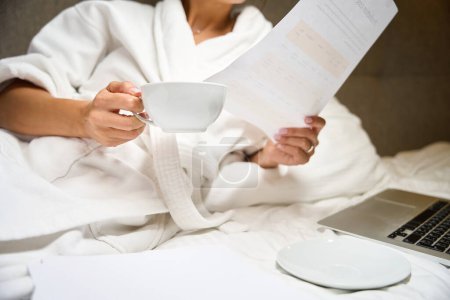 Photo for Cropped businesswoman wearing bathrobe with cup of tea or coffee watching document on bed in hotel room at morning time. Concept of business trip, vacation and travelling - Royalty Free Image
