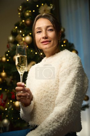 Téléchargez les photos : Focus sur l'avant-plan de la jeune belle femme caucasienne avec du champagne en verre en regardant la caméra pendant Noël ou le Nouvel An à la maison la nuit. Concept de vacances d'hiver célébrant - en image libre de droit