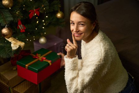 Téléchargez les photos : Vue en angle élevé de jeune femme caucasienne attrayante souriante mettant boîte cadeau sous l'arbre de Noël et montrant geste de silence pendant Noël ou Nouvel An à la maison la nuit. Vacances d'hiver célébrant - en image libre de droit