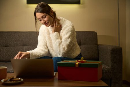 Photo for Young beautiful focused caucasian woman watching laptop while sitting on couch at table with gift box during Christmas or New Year at home at night time. Concept of winter holidays celebrating - Royalty Free Image