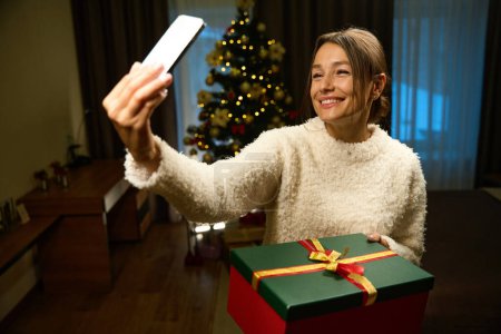 Téléchargez les photos : Focus sur le premier plan de heureuse femme caucasienne heureuse prenant selfie sur smartphone avec boîte cadeau pendant Noël ou Nouvel An à la maison floue avec arbre de Noël la nuit. Vacances d'hiver célébrant - en image libre de droit