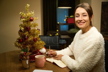 Téléchargez les photos : Jeune femme souriante jolie européenne regardant la caméra pendant l'écriture de ses souhaits ou notes dans un carnet à table pendant Noël ou le Nouvel An à la maison la nuit. Concept de vacances d'hiver célébrant - en image libre de droit