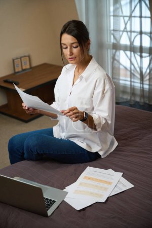 Photo for Young focused beautiful caucasian businesswoman watching documents on bed in hotel room at daytime. Concept of business trip, vacation and travelling - Royalty Free Image