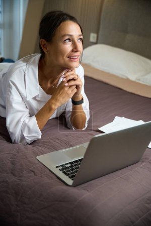 Téléchargez les photos : Jeune belle femme d'affaires caucasienne réfléchissant pendant le travail sur ordinateur portable sur le lit dans la chambre d'hôtel le jour. Concept de voyage d'affaires, vacances et voyages - en image libre de droit
