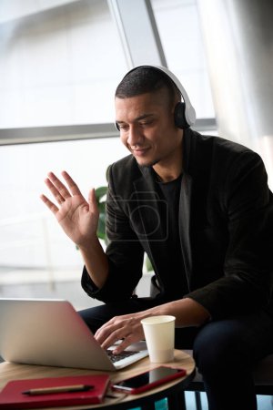 Photo for Smiling young male call center manager waving his hand to online interlocutor during videocall while sitting in front of laptop - Royalty Free Image