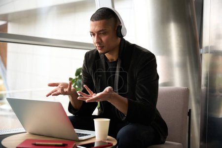 Foto de Calma consultor masculino sentado frente a la computadora portátil y gesticulando con las manos al tratar de explicar algo al interlocutor en línea - Imagen libre de derechos