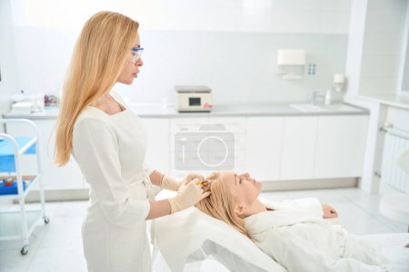 Photo for Self-confident woman cosmetologist in protective eyeglasses and gloves making injections into female scalp, hair plasmolifting procedure, improving of hair growth - Royalty Free Image