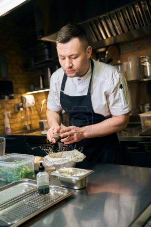 Téléchargez les photos : Chef caucasien concentré saupoudrer d'épices avec une passoire à thé dans une huître dans un nid à table au restaurant. Concept de délicieuse alimentation saine - en image libre de droit