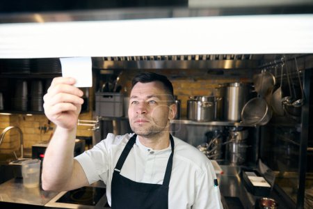 Téléchargez les photos : Chef caucasien professionnel portant une note de lecture uniforme dans un restaurant flou. Concept de saine alimentation savoureuse - en image libre de droit