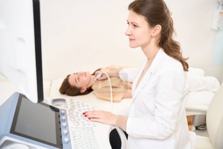 Foto de Médico caucásico femenino enfocado haciendo ecografía de la glándula tiroides de la mujer joven europea en la clínica. Concepto de asistencia sanitaria - Imagen libre de derechos