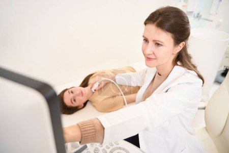 Téléchargez les photos : Médecin caucasien femme faisant une échographie de la glande thyroïde de la jeune femme européenne et ils regardent l'écran en clinique. Concept de soins de santé - en image libre de droit