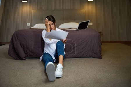 Photo for Young tired pretty caucasian businesswoman with headache sitting with documents on floor by bed in hotel room at daytime. Concept of business trip, vacation and travelling - Royalty Free Image