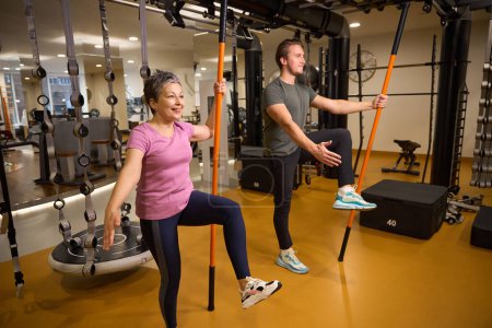 Photo for Male trainer teaching adult smiling caucasian woman wearing sportswear doing sport exercise with sports sticks in gym. Concept of modern healthy lifestyle - Royalty Free Image