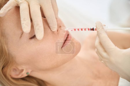 Téléchargez les photos : Dermatocosmétologue qualifié effectuant des microinjections de charge à base d'acide hyaluronique dans le nez du client féminin, le contouring du visage, la médecine esthétique - en image libre de droit