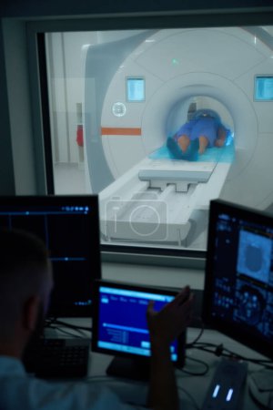 Radiographer seated at desk in control room operating MRI scanner using computer equipment while watching patient through viewing window