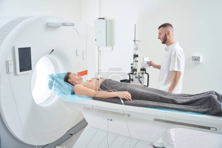 Radiologischer Techniker bedient Kontrastinjektor, während er Patient mit EKG-Elektroden auf Brust liegend auf CT-Tisch beobachtet
