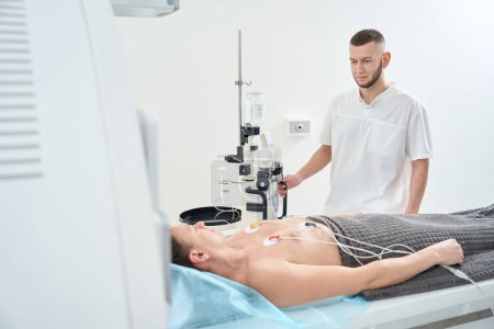 Radiographer standing near contrast injector while watching man with ECG electrodes placed on chest lying supine on computed tomography table