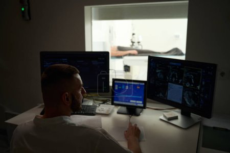 Radiologic technologist seated at desk in control room viewing patient CT scans on computer monitor