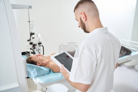 Röntgengerät mit Tablet-Computer neben Mann mit EKG-Elektroden auf Brust liegend auf CT-Tisch neben Kontrastinjektor