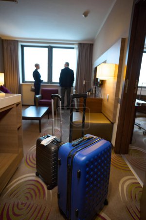 Focus on foreground of suitcases with blurred background of businessman and businesswoman standing and looking from window in hotel room at daytime. Business trip, vacation and travelling. Teamwork