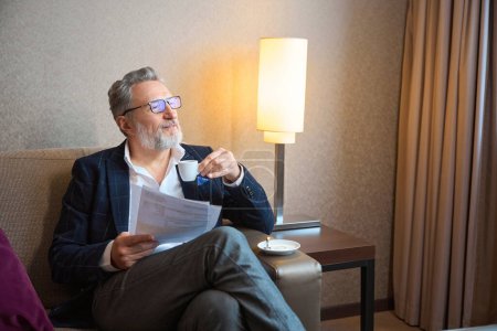 Téléchargez les photos : Homme d'affaires caucasien adulte concentré avec des papiers buvant du café dans une tasse et regardant loin sur le canapé dans la chambre d'hôtel pendant la journée. Concept de voyage d'affaires, vacances et voyages - en image libre de droit