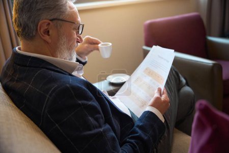 Foto de Empresario masculino caucásico adulto con taza de té o café viendo documento de papel en el sofá en la habitación de hotel durante el día. Concepto de viaje de negocios, vacaciones y viajes - Imagen libre de derechos