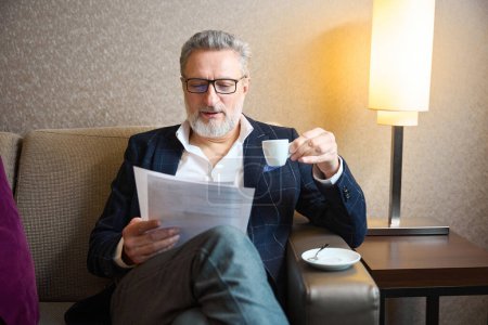 Foto de Hombre de negocios caucásico adulto enfocado con taza de té o café viendo documento de papel en el sofá en la habitación de hotel durante el día. Concepto de viaje de negocios, vacaciones y viajes - Imagen libre de derechos