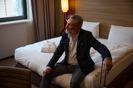 Téléchargez les photos : Homme entrepreneur caucasien adulte avec valise assise sur le lit et regardant loin dans la chambre d'hôtel pendant la journée. Concept de voyage d'affaires, vacances et voyages - en image libre de droit