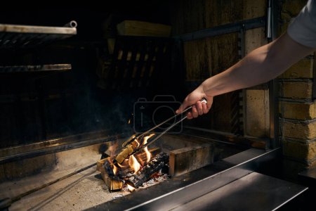 Photo for Male chef hand with kitchen tongs preparing woods in burning fire place for food cooking in restaurant. Concept of tasty healthy eating - Royalty Free Image