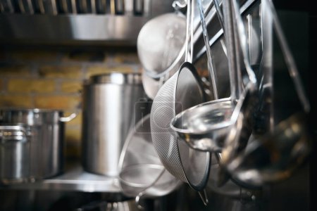 Photo for Focus on foreground of hanging cooking utensils and blurred table with saucepans on background in restaurant. Concept of tasty healthy eating - Royalty Free Image