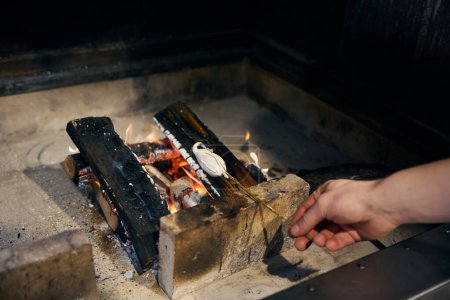 Photo for Cropped male chef hand frying marshmallow on tree branch in burning fire place in restaurant. Concept of delicious healthy eating - Royalty Free Image