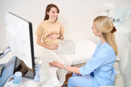 Téléchargez les photos : Gynécologue féminine préparer machine à échographie et en regardant jeune femme européenne souriante enceinte à la clinique. Concept de grossesse et de maternité - en image libre de droit