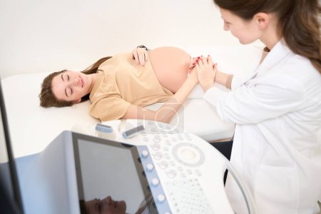 Foto de Ginecóloga y joven mujer embarazada europea cogidas de la mano y mirándose antes de la ecografía en la clínica. Concepto de embarazo y maternidad - Imagen libre de derechos