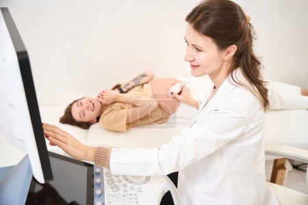 Téléchargez les photos : Gynécologue caucasienne effectuant une échographie du ventre enceinte de la jeune femme européenne floue heureuse à la clinique. Concept de grossesse et de maternité - en image libre de droit