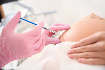 Foto de Enfoque selectivo de ginecóloga femenina inyectable con jeringa en vientre embarazada de mujer en clínica borrosa. Concepto de embarazo y maternidad - Imagen libre de derechos