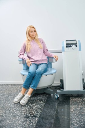 Photo for Satisfied blond woman sitting on cozy portable electromagnetic stimulation chair, effective therapy for problems in the lower back area that stimulating muscles and accelerates circulation - Royalty Free Image
