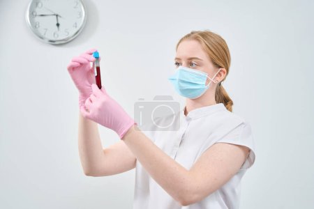 Trabajadora de laboratorio cualificada en mascarilla protectora comprobando la vacutainer con muestra de sangre antes de realizar investigación bioquímica, chequeo de salud