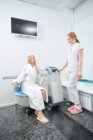 Trabajadora médica calificada comprobando el bienestar de la mujer que toma terapia de músculos del suelo pélvico sentada en una silla de estimulación electromagnética, clínica de medicina estética