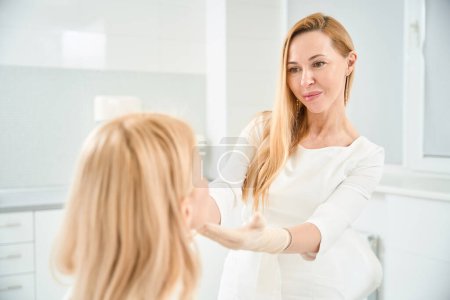 Photo for Attractive cosmetician looking at face of female client to find imperfects of skin she would be changing and improving during therapy, aesthetic medicine clinic - Royalty Free Image