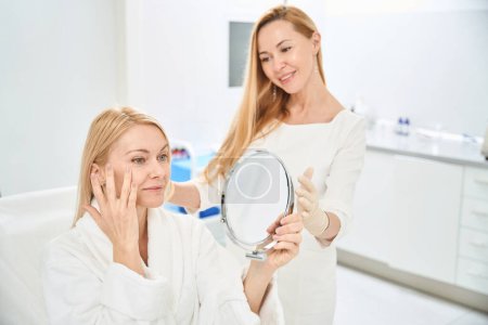 Lächelnde Kundin der Klinik für ästhetische Medizin zufrieden mit Narbenentfernung und Hautstraffung um die Augen, Blick auf ihr Spiegelbild im Spiegel, guter Service
