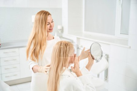 Téléchargez les photos : Cosmetologue tenant le miroir devant la cliente, regardant la réduction des cercles bleus et le gonflement sur son visage et sous les yeux après la procédure d'hydratation - en image libre de droit