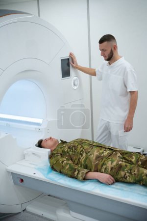 Foto de Tecnólogo apoyado en el pórtico del escáner de resonancia magnética mientras observa al paciente militar acostado en la mesa de exploración - Imagen libre de derechos