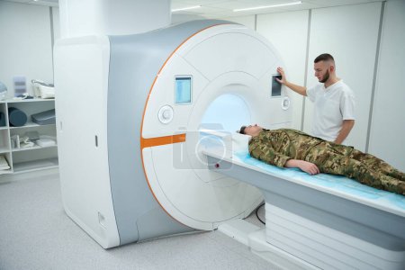Mediziner drückt Taste auf MRT-Gerät Portalsteuerung, während Militärpatient auf Scan-Tisch liegt