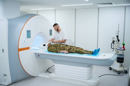 Photo for Serviceman lying supine on MRI table while healthcare professional placing plastic coil around his head - Royalty Free Image