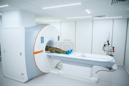 Photo for Serviceman in camouflage uniform lying supine on MRI table in scanning room - Royalty Free Image