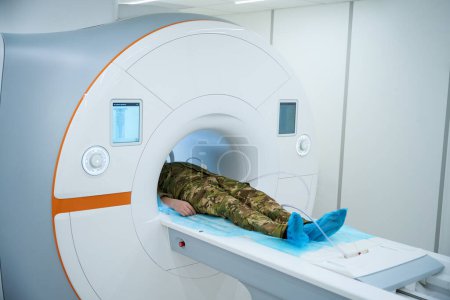 Photo for Military patient in camouflage uniform lying on his back on MRI table in scanning room - Royalty Free Image