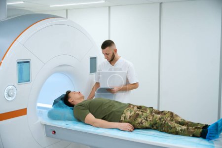 Technologist placing shoulder coil over arm of serviceman positioned on MRI table